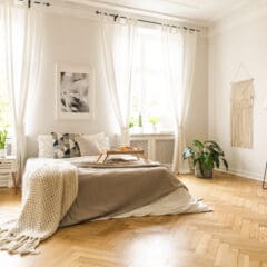 Bright bedroom with a large bed topped with neutral bedding, a knit blanket, and a breakfast tray. White curtains, plants, wall art, and macramé decor add to the cozy and airy atmosphere. Wooden herringbone floor.