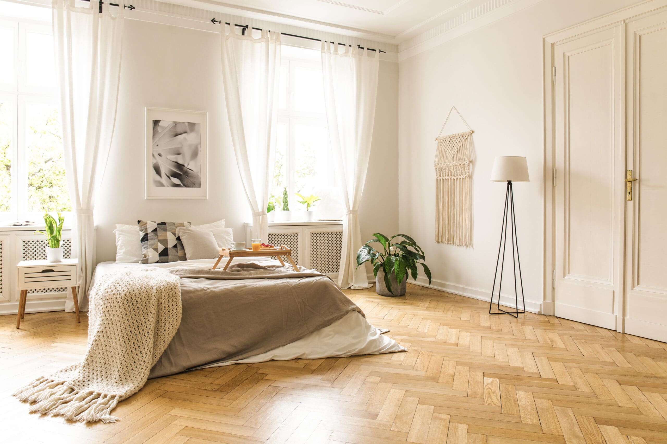 A bright, cozy bedroom with a neatly made bed adorned with pillows and a beige blanket. Sunlight streams through sheer white curtains. The room features a potted plant, wall art, and a standing lamp on herringbone wood flooring.