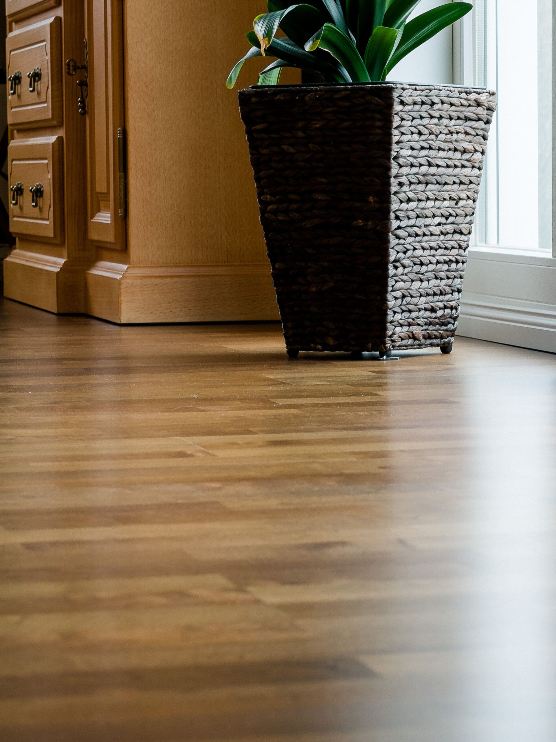 A wooden floor with a large, woven plant pot holding a leafy green plant. The pot is positioned near a wooden cabinet and a large window, allowing natural light to illuminate the room.