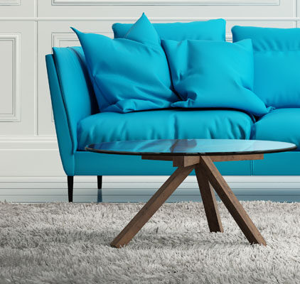 A modern living room with a bright blue sofa featuring several cushions. In front of the sofa is a round glass coffee table with wooden legs, placed on a textured gray carpet. The walls are white with paneled detailing.