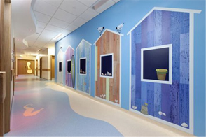 A brightly decorated hallway with walls painted to resemble colorful beach huts featuring empty blackboard windows. The floor has a wave-like pattern, and a pail is placed on one window ledge, adding to the seaside theme.