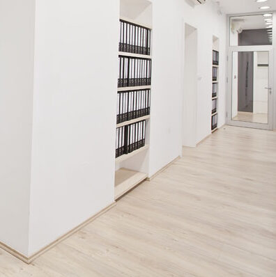 Minimalist office hallway with light wood flooring and white walls. Built-in shelves on the left hold organized black binders. A large mirror hangs on the right wall, reflecting the bright, well-lit space.