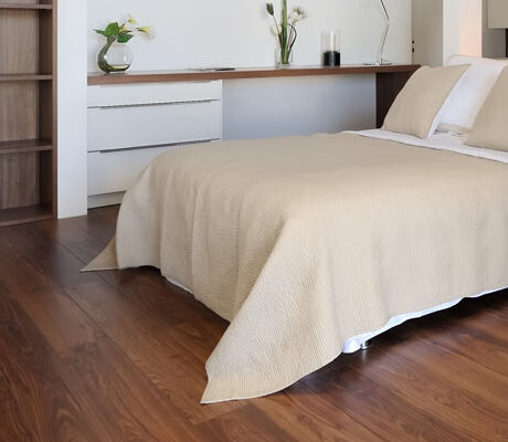 A minimalist bedroom with a neatly made bed featuring beige bedding. The room has a wooden floor, a built-in desk against the wall with a plant and candles, and shelving for storage. Soft natural light enhances the serene ambiance.