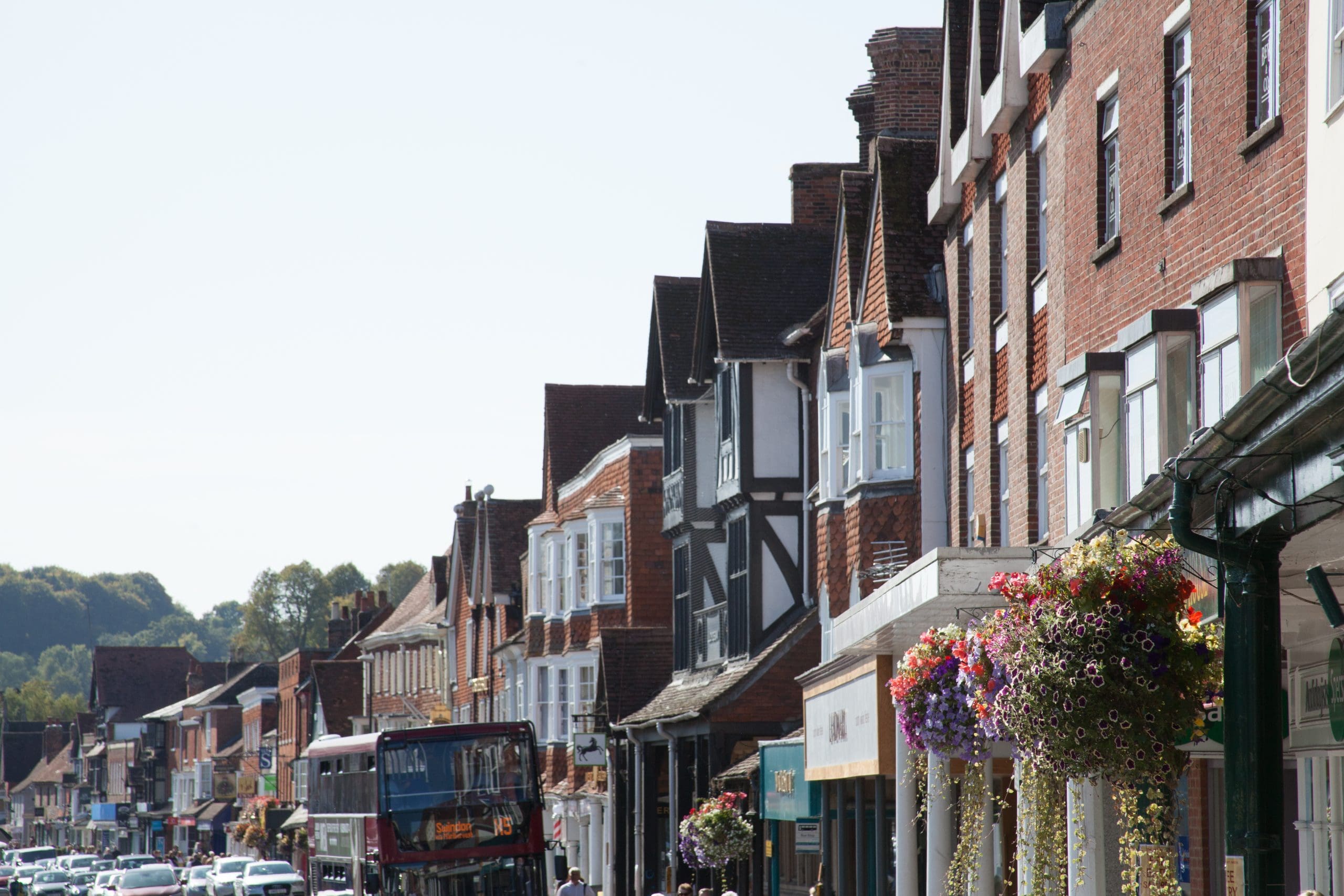 A row of traditional brick buildings lines a bustling street. There's a double-decker bus in the foreground, and colorful hanging flower baskets adorn the facades. The scenic background includes distant hills and a bright sky.