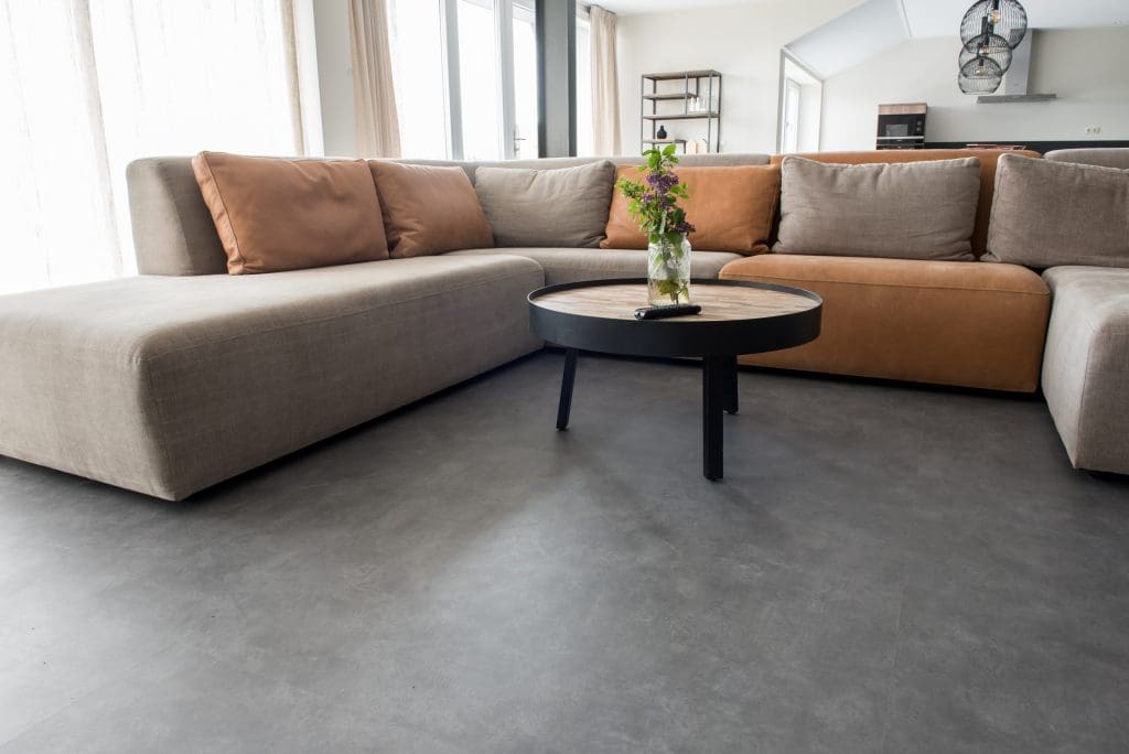 A modern living room with a large L-shaped sectional sofa featuring beige and brown cushions. A round wooden coffee table with a black base sits on a gray floor, with a vase of flowers on top. Natural light filters through large windows.