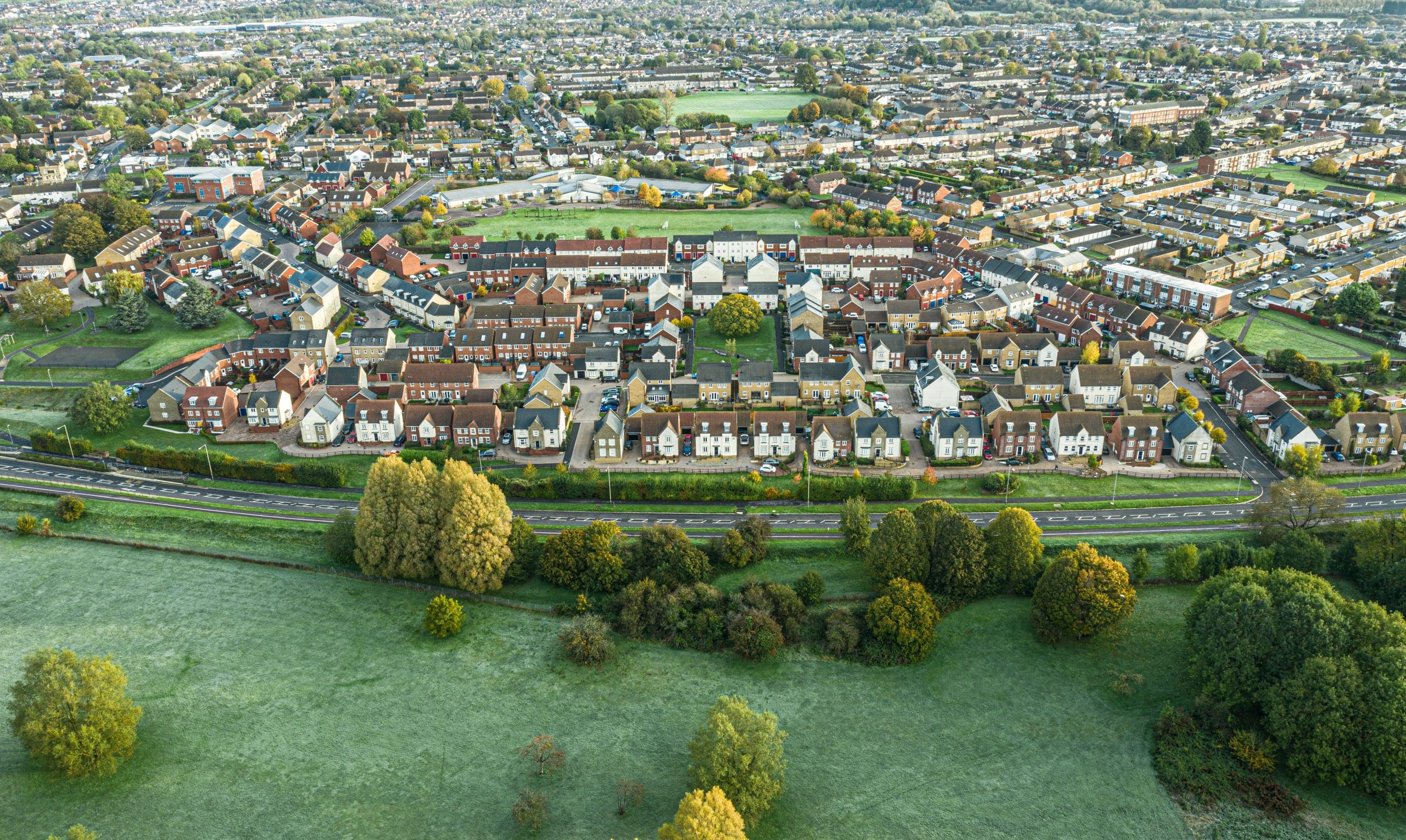 Aerial view of a suburban neighborhood with rows of houses, surrounded by green fields and trees. A railway line runs parallel to the edge of the residential area, separating it from the open landscape. The sky is clear.