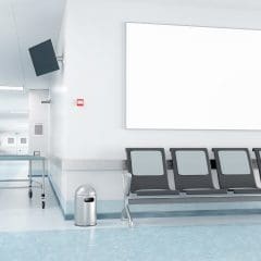 A modern hospital corridor with light blue flooring, gray-blue walls, and bright lighting. There's a row of four gray chairs, an empty stretcher trolley, a small trash can, a wall-mounted TV, and a large blank white board on the wall.