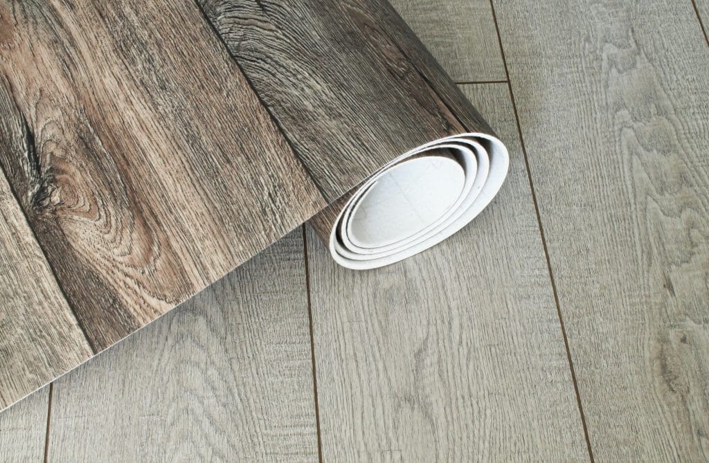 A roll of wood-patterned vinyl flooring is partially unrolled on a wooden floor. The vinyl displays a detailed brown wood grain, contrasting with the lighter gray wood texture of the existing floor.