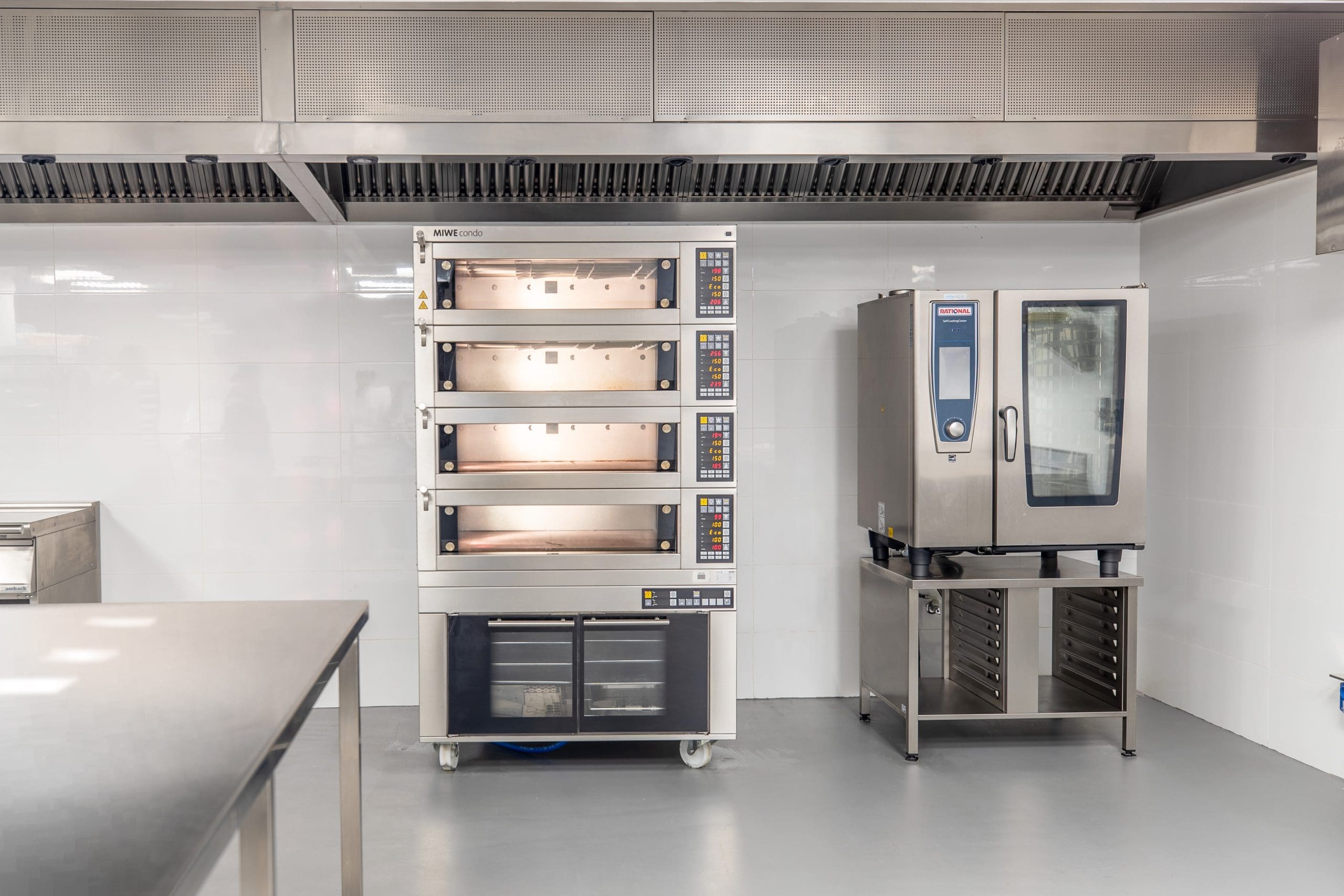 A clean, modern commercial kitchen with a stainless steel double rack oven on the left and a combi oven on the right. The ovens are installed under a large ventilation hood against a tiled white wall. The floor is smooth and grey.