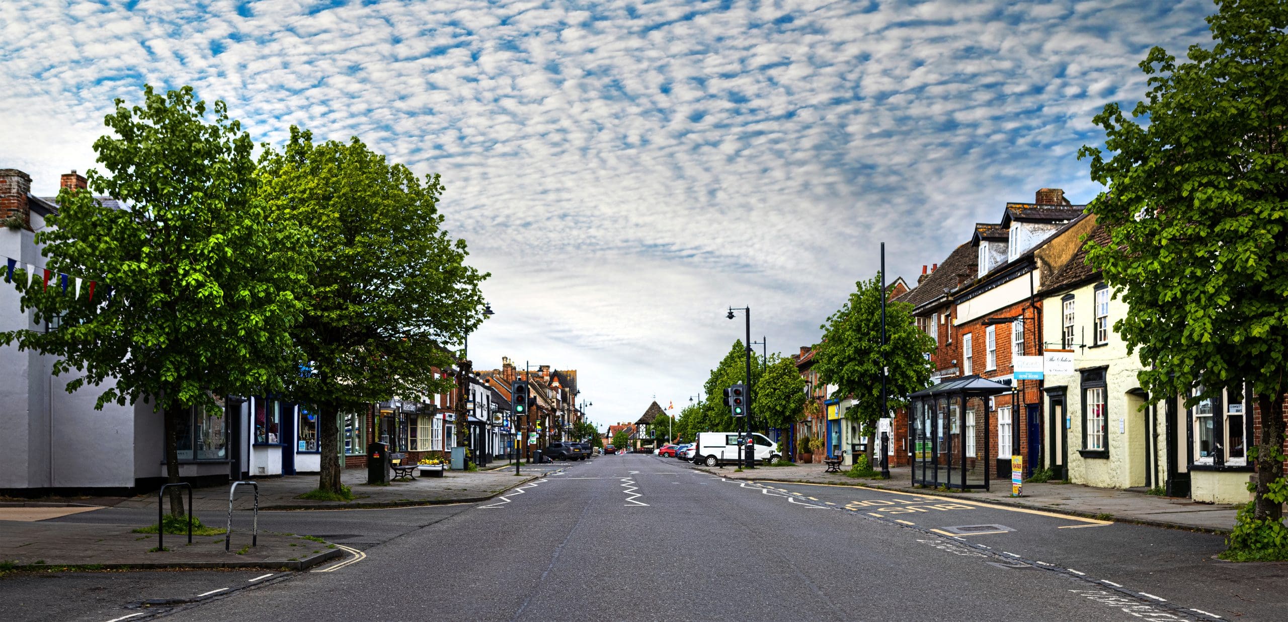 A picturesque small town street with shops and buildings lining both sides. The road is empty, and trees are scattered along the sidewalks. The sky above is filled with patterned clouds, creating a serene atmosphere.