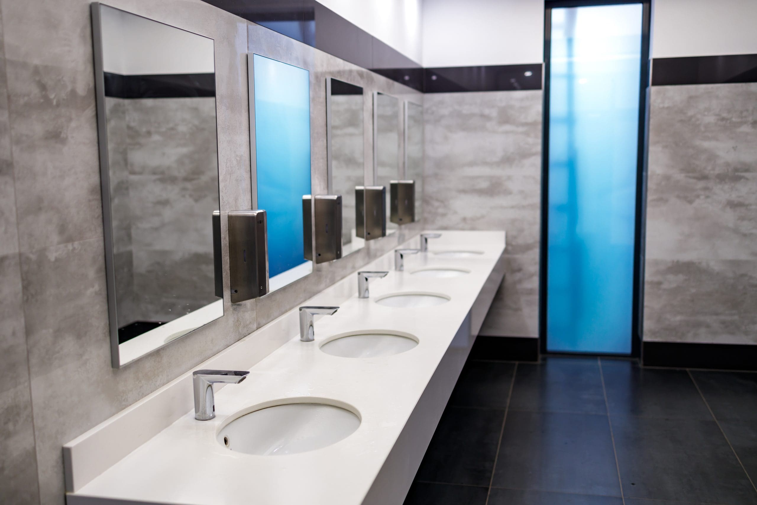 A modern restroom with a long white counter featuring five sinks and mirrors. Each sink has a silver faucet and a soap dispenser mounted on the wall. The room has gray tiled walls and a blue glass door in the background.