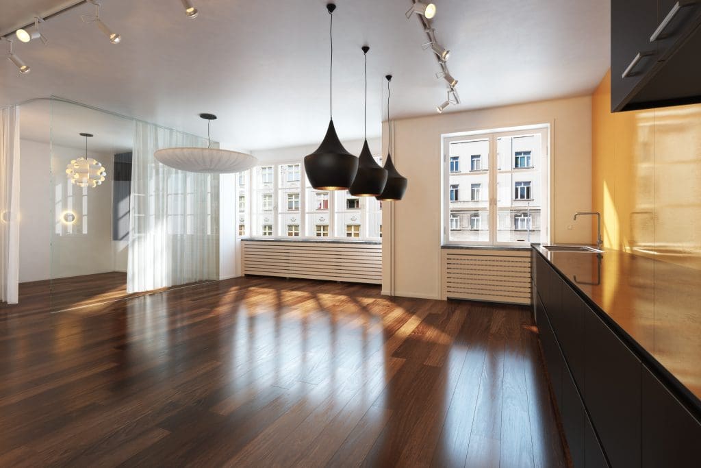 A spacious, modern kitchen with dark wooden floors and ceiling spotlights. It features sleek black cabinets, pendant lights, and large windows with a view of neighboring buildings. A glass partition separates a bright, open dining area.