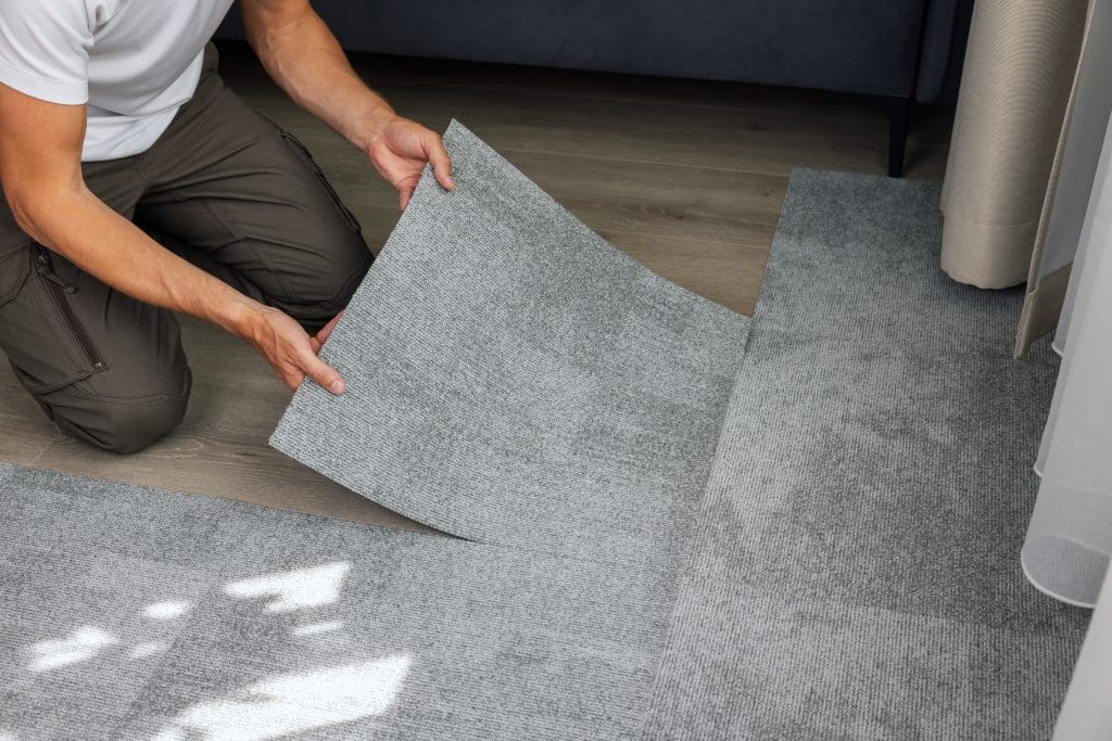 A person in a white shirt and dark pants is installing gray carpet tiles on a wooden floor. The person is kneeling and fitting the tiles together, with natural light casting shadows on the floor.