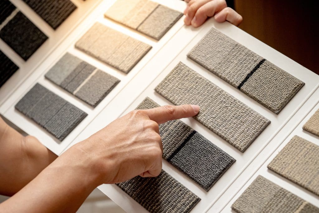 A person points to a fabric sample on a board displaying various textured fabric swatches in neutral tones. The samples range from light beige to dark gray.