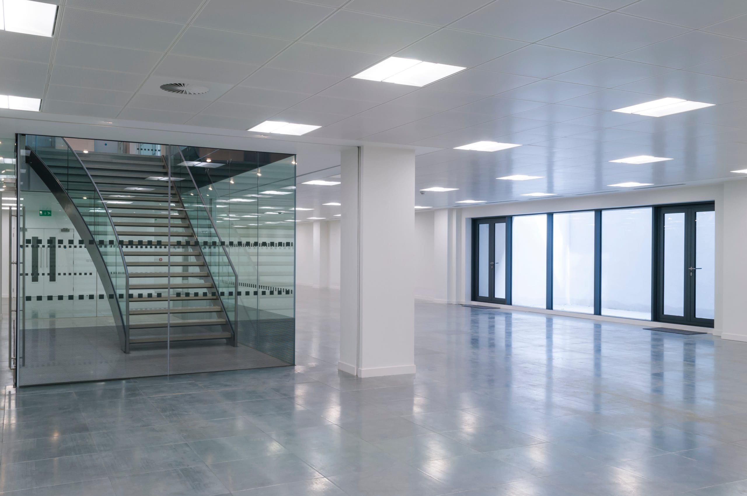 An empty, modern office space with large windows. The room features a glass-walled staircase, glossy light gray floor tiles, and ceiling lights. The area has a minimalist design with ample natural and artificial lighting.