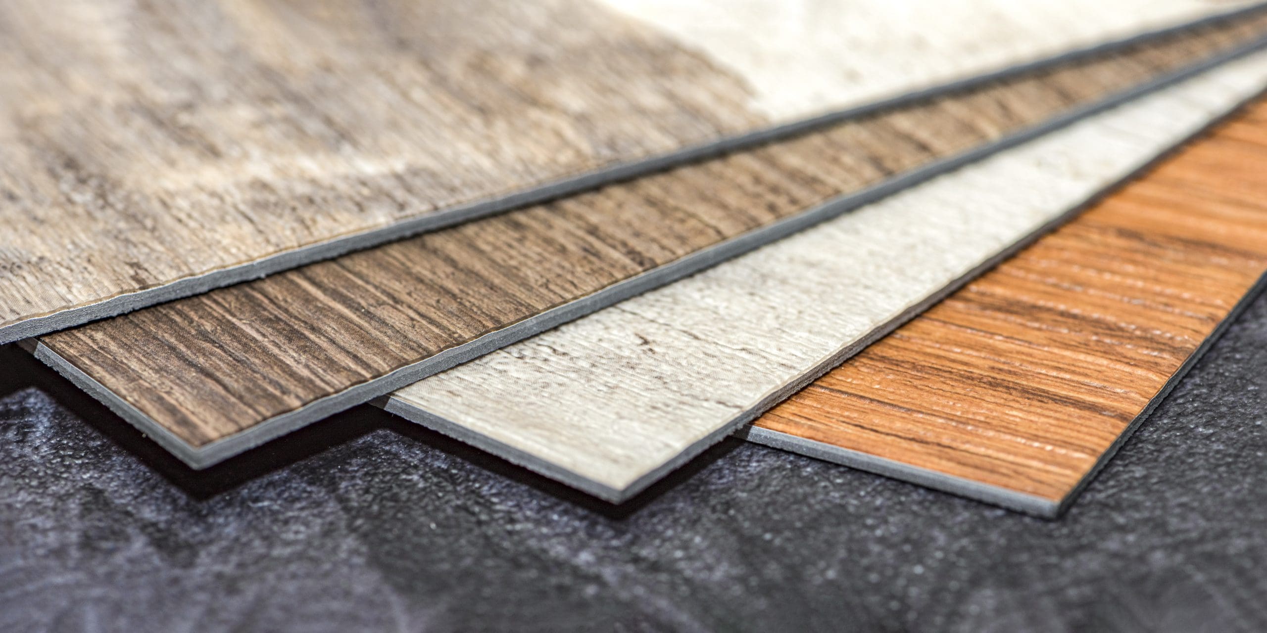 Close-up of four different laminate flooring samples in various wood and tile textures, stacked in a fan shape. The samples feature colors ranging from dark brown to light tan. The background is a textured dark surface.