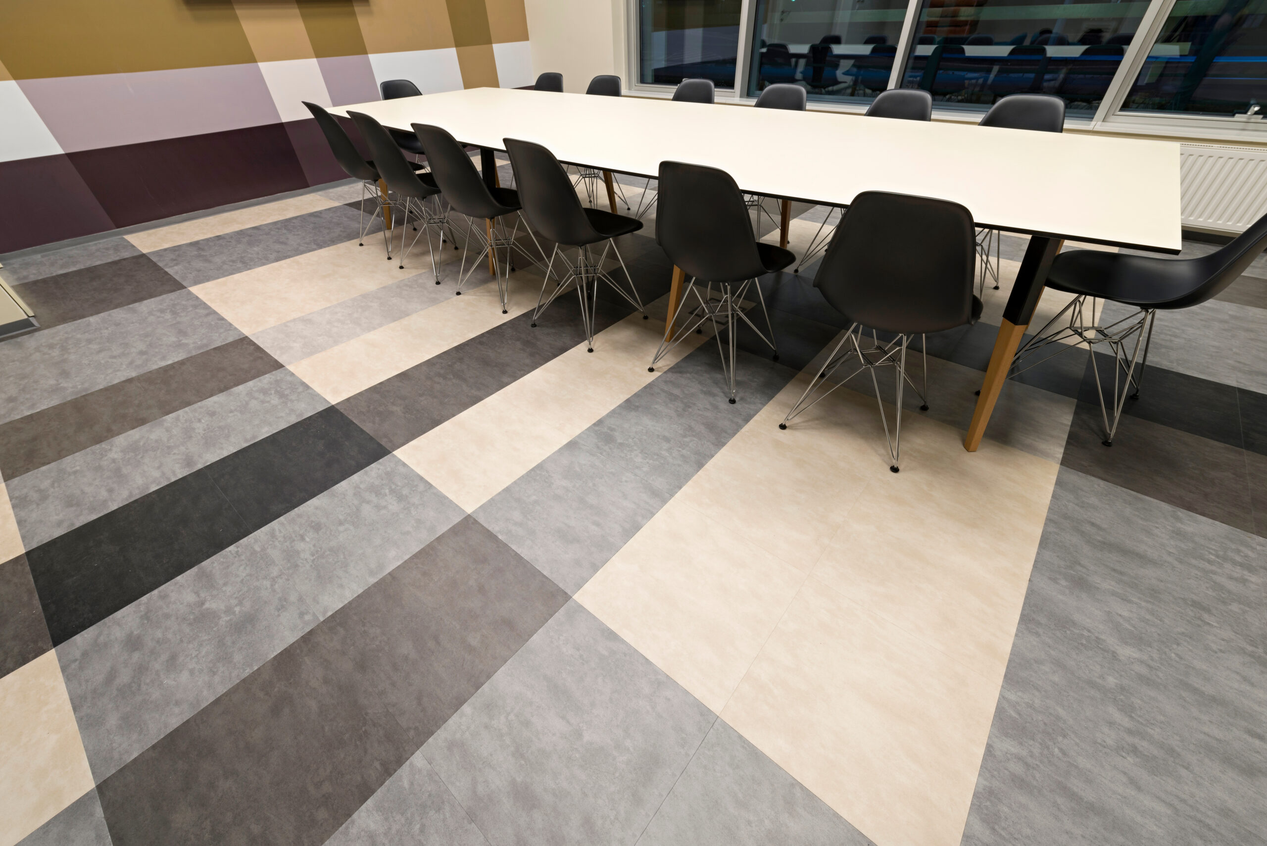 A modern conference room with a long white table surrounded by black chairs. The floor features a bold, geometric pattern in shades of grey, black, and beige. Large windows line one wall, letting in natural light.