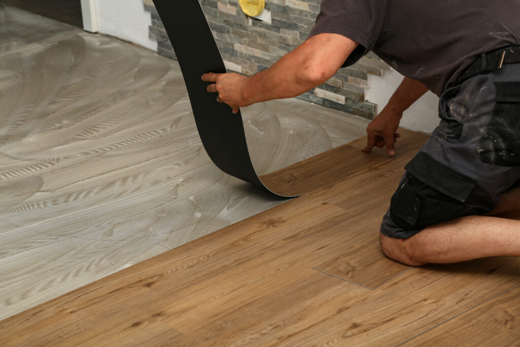 A person installs vinyl wood-look flooring, kneeling on the ground and carefully placing a plank onto adhesive spread on the floor near a wall with stone tile accents.