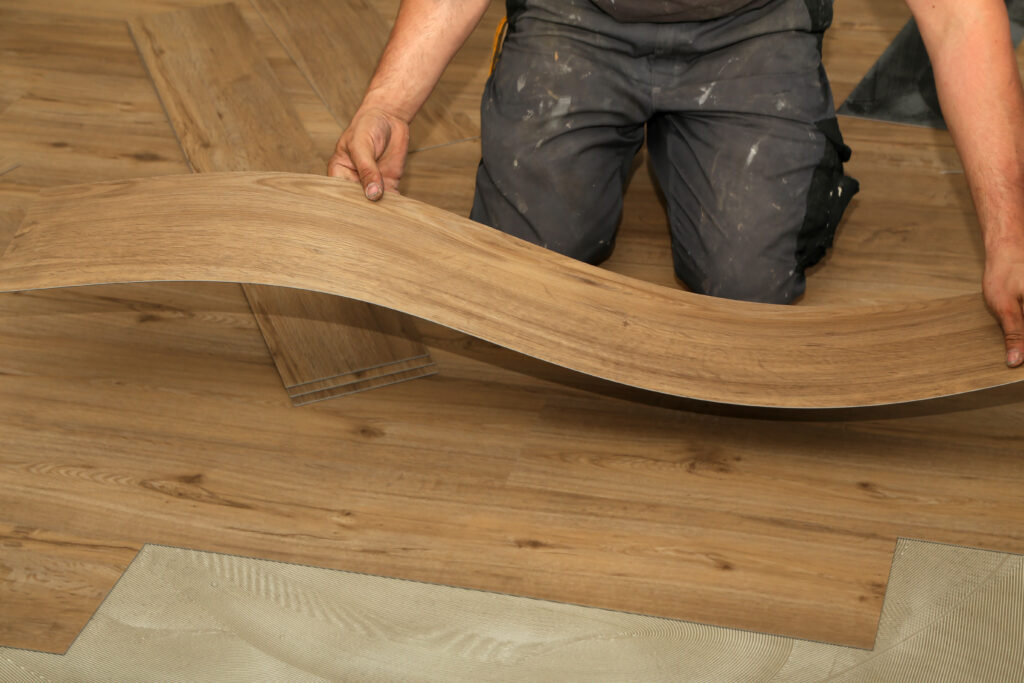 A person installs vinyl plank flooring, holding a flexible plank above an unfinished floor with adhesive and other planks visible.