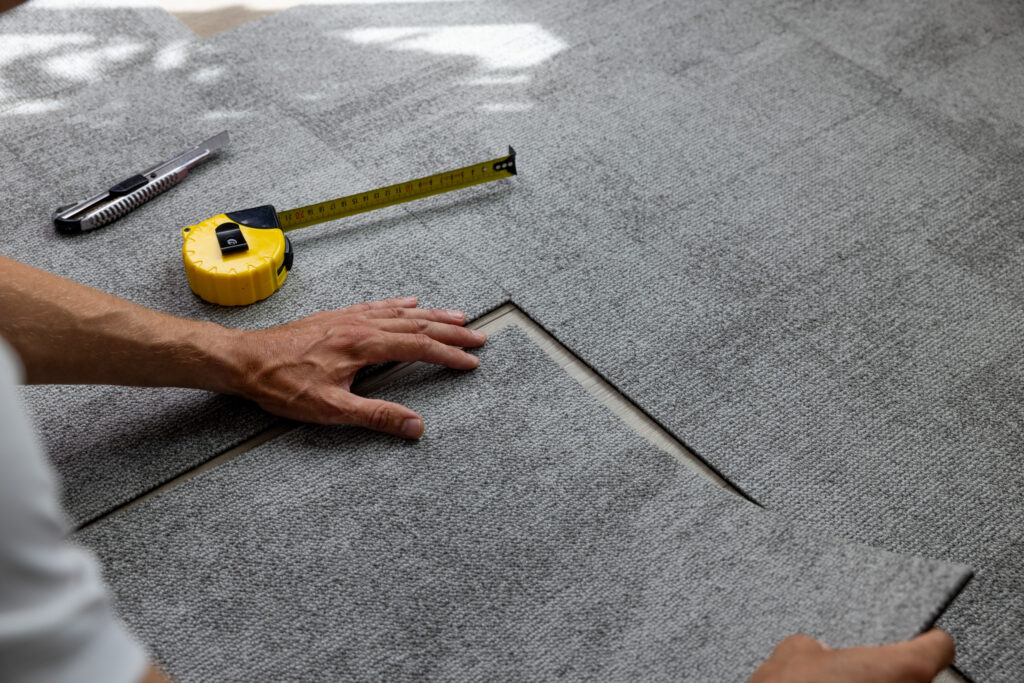 A person installs grey carpet tiles, aligning them precisely. A yellow measuring tape and a utility knife are placed nearby on the carpet surface. Sunlight partially illuminates the workspace.